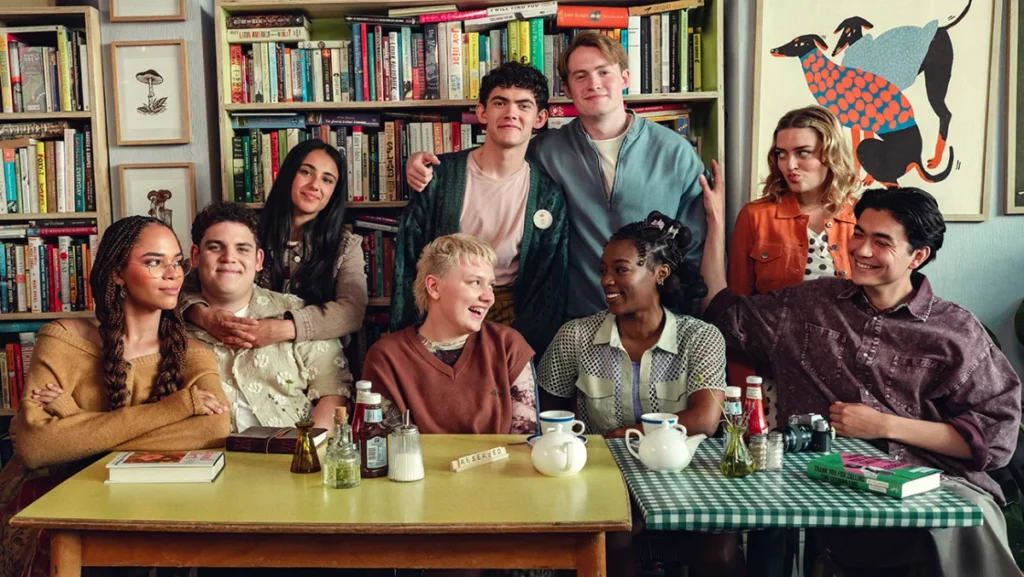 "Nine young adults sit and stand around a table in a cozy cafe or library, surrounded by bookshelves and artwork, enjoying tea and conversation, with condiments, teapots, and cups visible on the tables."