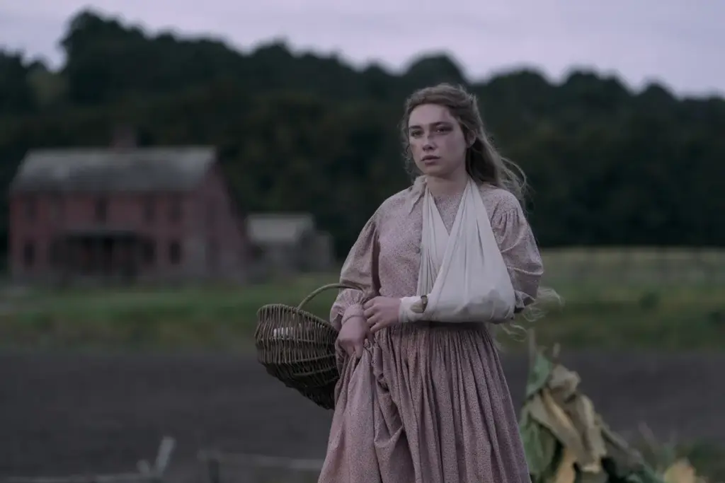 "Young woman in historical dress with arm in a sling stands in a rural field, holding a basket, with a rustic farmhouse and trees in the background under a cloudy sky."