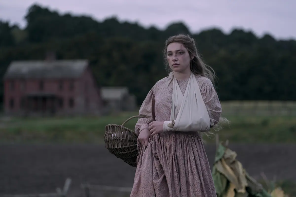 "Young woman in historical dress with arm in a sling stands in a rural field, holding a basket, with a rustic farmhouse and trees in the background under a cloudy sky."
