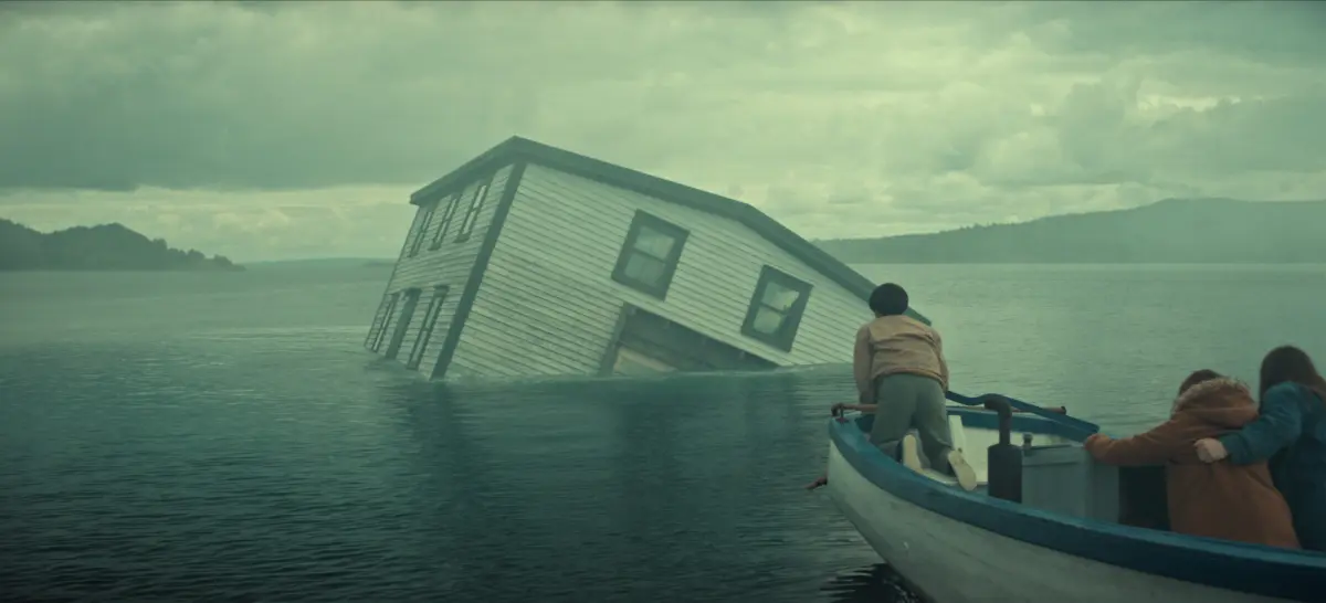 "A partially submerged house tilts in a misty lake as three people in a small boat observe, with cloudy skies and distant hills creating a dramatic, mysterious atmosphere."